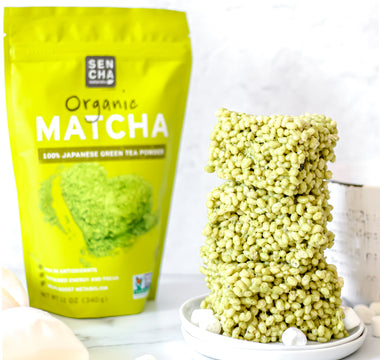 Photo of a bag of Sencha Naturals Matcha green tea powder next to a stack of 3 rice krispies treats squares on a white plate, with a few small marshmallows strewn about, all on a white background