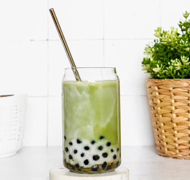 Photo of a clear glass jar containing boba bubble tea with a silver straw, with a small green plant in the back, all on a white background