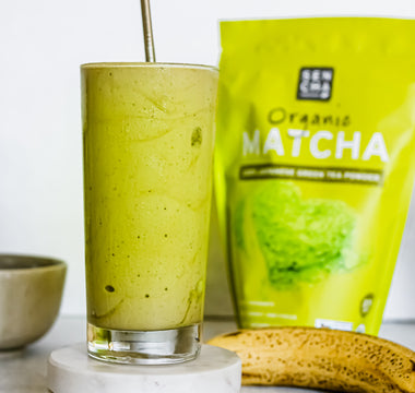 Photo of a bag of sencha naturals organic japanese green tea powder on the right, with a glass of a green tea milkshake on the left, a banana in between, all set on a white background