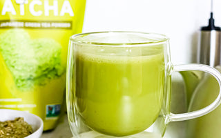 Photo of a bag of Sencha Naturals organic Japanese matcha green tea powder, with a clear mug in the foreground containing a matcha green tea latte, as well as a small electric whisk in the back, all on a white background