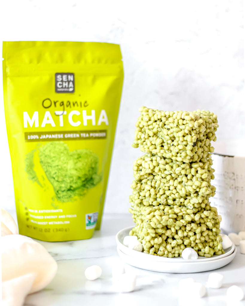 Photo of a bag of Sencha Naturals Matcha green tea powder next to a stack of 3 rice krispies treats squares on a white plate, with a few small marshmallows strewn about, all on a white background