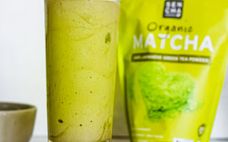 Photo of a bag of sencha naturals organic japanese green tea powder on the right, with a glass of a green tea milkshake on the left, a banana in between, all set on a white background