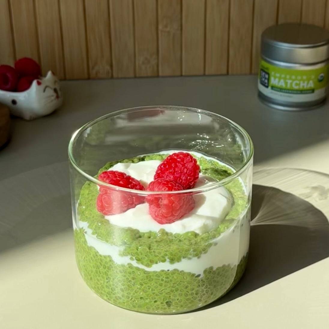 A glass cup filled with layered matcha chia pudding and yogurt, topped with three fresh raspberries. A tin of organic matcha powder sits in the background on a sunlit counter