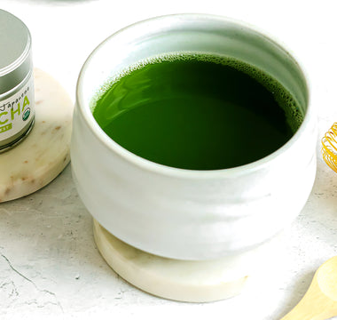 Photo of a small silver tin of Sencha Naturals Ceremonial Grade Matcha Powder on the left, a small white cup of liquid green tea in the center, and a small wooden spoon and bamboo whisk on the right, all on a white background