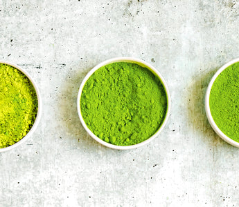 Photo of 3 small bowls on a grayish-white background, each containing matcha green tea powder in varying shades from light green on the left to dark green in the center.