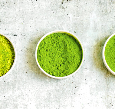 Photo of 3 small bowls on a grayish-white background, each containing matcha green tea powder in varying shades from light green on the left to dark green in the center.