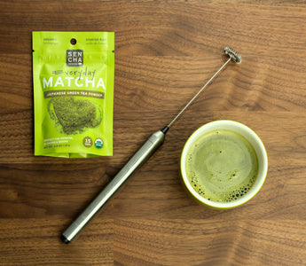 Photo of a bag of Sencha Naturals matcha green tea powder on the left, a small white cup of liquid green tea on the right, and a small electric whisk in the middle, all on a wooden table