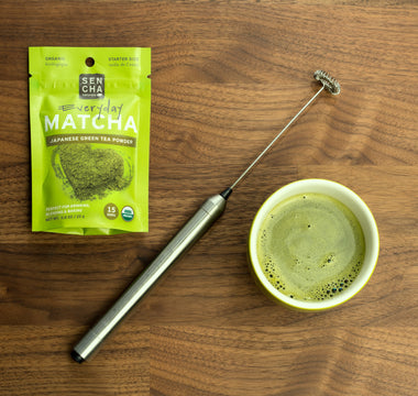 Photo of a bag of Sencha Naturals matcha green tea powder on the left, a small white cup of liquid green tea on the right, and a small electric whisk in the middle, all on a wooden table