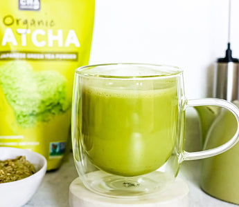 Photo of a bag of Sencha Naturals organic Japanese matcha green tea powder, with a clear mug in the foreground containing a matcha green tea latte, as well as a small electric whisk in the back, all on a white background