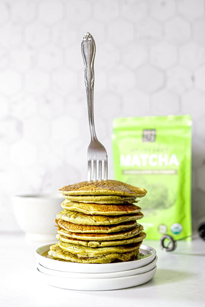 Photo of a stack of pancakes made with matcha green tea powder, with a silver fork stuck on top, and a bag of matcha green tea in the background, all set on a white countertop.