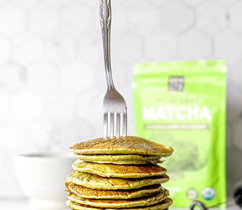Photo of a stack of pancakes made with matcha green tea powder, with a silver fork stuck on top, and a bag of matcha green tea in the background, all set on a white countertop.