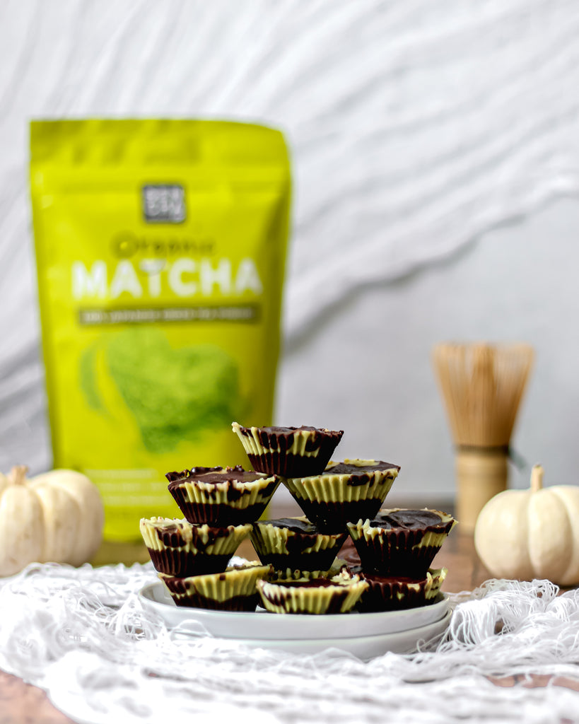 Photo of Sencha naturals matcha green tea powder in a bag, a stack of chocolate matcha dessert cups in the foreground, as well as a bamboo whisk in the back along with some small white decorative pumpkins, all on a white background