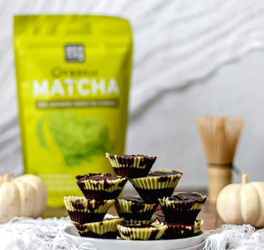 Photo of Sencha naturals matcha green tea powder in a bag, a stack of chocolate matcha dessert cups in the foreground, as well as a bamboo whisk in the back along with some small white decorative pumpkins, all on a white background
